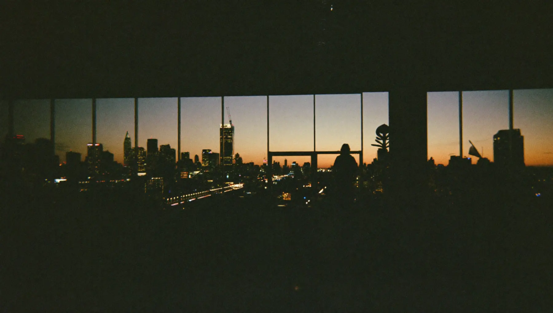 The city skyline seen through the club's floor to ceiling glass before dawn, a lone figure at the window